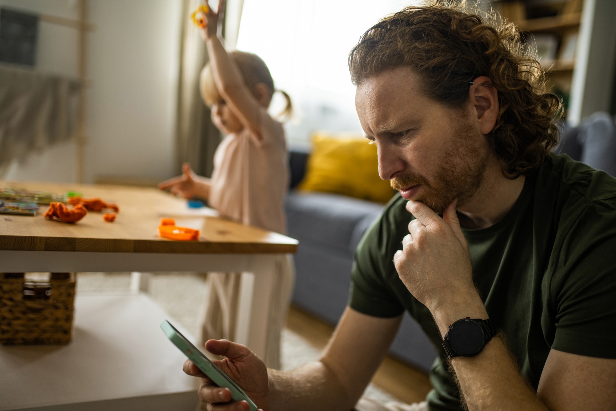 Worried man using phone while his little daughter is playing at home